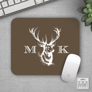 May include: Brown mouse pad with a white deer head and antlers, and the letters "M K" on either side of the deer. The mouse pad is on a gray surface.