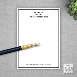 May include: White notepad with a black border and a black and white illustration of eyeglasses at the top. The text "THOMAS FITZGERALD" is printed below the illustration. The text "THOMASFITZGERALD@GMAIL.COM - 212-422-2029" is printed at the bottom of the notepad. A black and gold pen is lying on top of the notepad. The text "THE GENTLEMAN'S STUDY" is printed in a black square with a white background and a small mustache icon at the top.