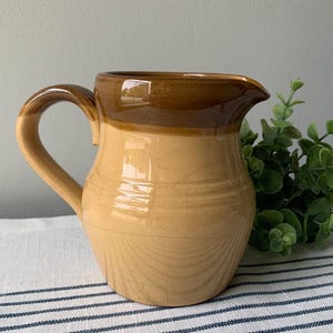 May include: A two-toned ceramic pitcher with a handle and spout. The top is a dark brown, transitioning to a light tan base. The pitcher has a crackled glaze and is likely vintage. The pitcher is on a striped surface.