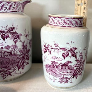 May include: White ceramic pitcher and jar set with a burgundy landscape and leaf design. The pitcher has a spout and handle, while the jar has a lid. Both pieces feature a detailed, vintage aesthetic.