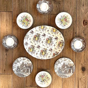 May include: An assortment of vintage china on a wooden surface. The set includes saucers with gold rims, floral-patterned plates, and a large oval platter with a fruit design. Some plates feature a black and white landscape scene.