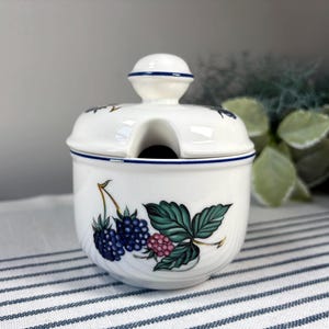 May include: White ceramic sugar bowl with a lid and a small handle. The bowl is decorated with a design of blackberries and green leaves. The rim and handle are trimmed with a blue line.