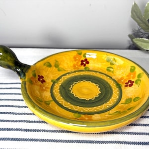 May include: A round ceramic dish with a single handle, featuring a vibrant yellow base color. The interior is decorated with a central dark green circle, surrounded by painted orange and red floral motifs and green leaves on a yellow background.
