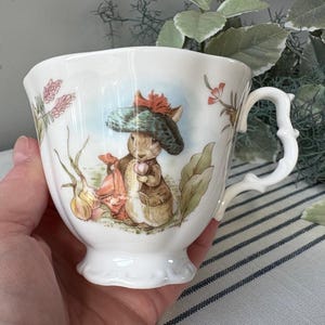 May include: White teacup with a detailed illustration of a rabbit wearing a hat and holding a pink object. The teacup has a decorative handle and floral accents. The cup is held in a hand.