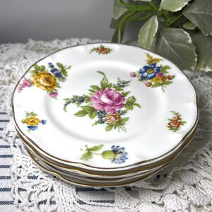 May include: A stack of white porcelain plates with scalloped edges and gold trim. Each plate features a central bouquet of pink roses surrounded by smaller clusters of yellow, blue, and orange flowers with green leaves, arranged in a floral pattern.