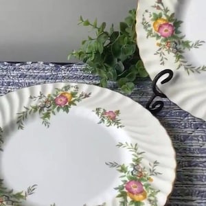 May include: Two white porcelain plates with a floral design. The plates have a scalloped edge and a gold rim. The floral design includes pink, yellow, and green elements. One plate is on a black metal stand.