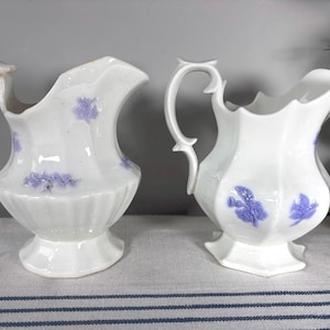 May include: Two white ceramic pitchers with a floral design in light purple. The pitchers have a handle and a spout. The pitchers are on a table with a blue and white striped cloth.