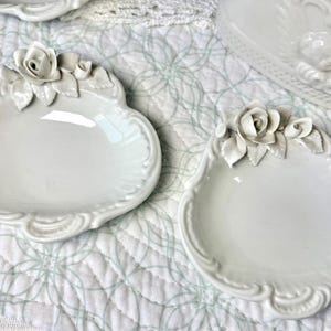 May include: Two white ceramic decorative dishes with a rose and leaf design. The dishes have a scalloped edge and are placed on a white quilted surface. The dishes are likely for decorative purposes.