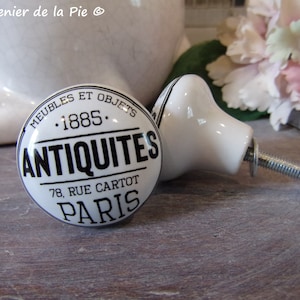 May include: Two white ceramic cabinet knobs with black text. One round knob reads "ANTIQUITES PARIS" and "1885". The other knob is teardrop-shaped. Both knobs have metal hardware attached for mounting on furniture.