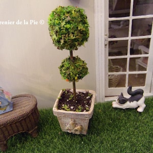 May include: A miniature topiary tree with two spherical green foliage sections in a square, aged-looking pot. A small, decorative table and a black and white cat figurine are nearby. The scene is set on artificial grass.