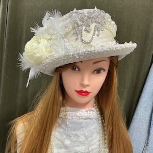 May include: A cream-coloured hat featuring a wide brim and a textured crown. The hat is adorned with a large cream rose, white feathers, strands of white pearls, and a prominent silver-toned decorative brooch with clear crystals and hanging chains.