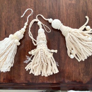 May include: Three white tassel ornaments with pearl accents. The tassels are made of a combination of fabric and rope.