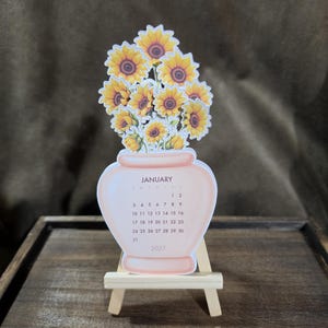 May include: A die-cut paper calendar for January 2027 shaped like a light peach vase filled with yellow sunflowers. The calendar displays the month name, days of the week, and dates. It is displayed on a small, light-colored wooden easel.
