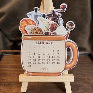 May include: A die-cut paper calendar for January 2027 shaped like a coffee mug, displayed on a small wooden easel. The mug features illustrations of a croissant, cookie, coffee maker, creamer, and whipped cream. The calendar grid shows dates 1-31.