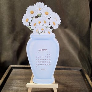 May include: A die-cut paper calendar shaped like a light blue vase filled with white daisies. The vase features the text JANUARY 2027 and a monthly calendar grid. The item is displayed on a small, light-colored wooden easel stand.
