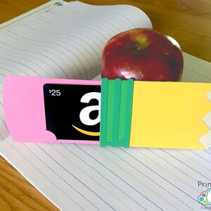 May include: A printable craft project for making a pencil-shaped gift card holder. The holder is made of yellow, pink, and green paper and has a slot for a gift card. The gift card shown is a $25 Amazon gift card. The project is titled "Printable Cuttable Creatables".