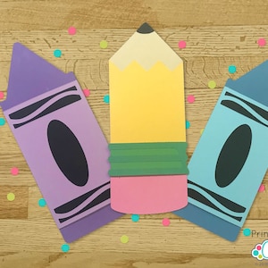 May include: Three printable cut-out shapes of school supplies: a purple crayon, a yellow pencil, and a blue crayon. The shapes are on a woodgrain background with colourful confetti.