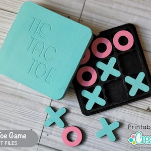 May include: A teal blue Tic Tac Toe game with a black game board and pink and teal game pieces. The game board is labeled "Tic Tac Toe". The game pieces are circles and X's. The game is made of wood and is laser cut.