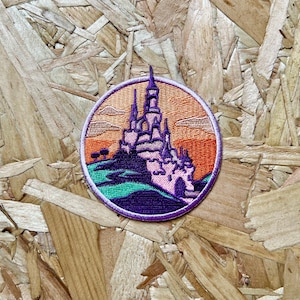 Könnte beinhalten: Ein gestickter Aufnäher in Lila und Rosa, der ein Schloss mit Sonnenuntergang im Hintergrund zeigt. Das Schloss hat mehrere Türme und ein Tor. Der Aufnäher ist rund und hat einen weißen Umriss.