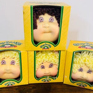 May include: Four Cabbage Patch Kids ear muffs in their original yellow boxes. The ear muffs feature the doll's face with different hair colors: brown and yellow. The boxes have the Cabbage Patch Kids logo.