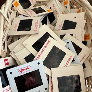 May include: A wicker basket overflowing with vintage film slides. The slides, labeled with brands like Kodachrome and Anscochrome, feature black rectangular windows. The slides are in various states of disarray, creating a nostalgic aesthetic.