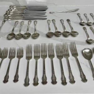 May include: A collection of antique silverware, including forks, spoons, and knives, arranged on a white surface. The silverware has ornate detailing and a silver finish, perfect for formal dining. The set includes various serving utensils.