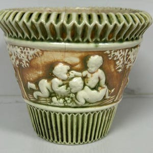 May include: A green and brown ceramic planter with a raised design of two cherubs playing with grapes. The planter has a scalloped rim and a ribbed base.