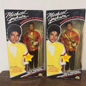 May include: Two boxed Michael Jackson Superstar of the 80's dolls. Each box features an image of Michael Jackson in a yellow vest and a red jacket. The boxes include a glittering glove, microphone, and posing stand. The text reads "Authentic Stage Outfit."