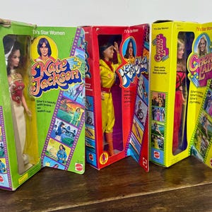 May include: Three vintage Mattel "TV's Star Women" dolls in their original boxes. The boxes feature images of Kate Jackson, Cheryl Ladd, and another woman. The dolls are dressed in various outfits, including a yellow jumpsuit and a red dress. The boxes are green, red, and yellow.