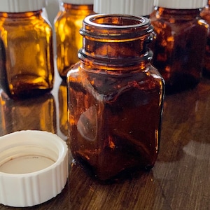 May include: A group of six amber glass bottles with white lids. The bottles are arranged in a row on a wooden surface. The bottles are all the same size and shape, and they have a square base.