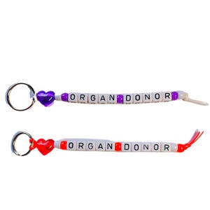 May include: Two keychains with the words "ORGAN DONOR" spelled out in white beads. The top keychain has a purple heart charm and the bottom keychain has a red heart charm.