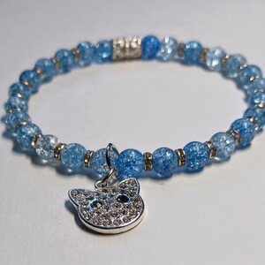 May include: A blue crystal beaded bracelet with a silver charm in the shape of a cat's head. The charm is encrusted with rhinestones.
