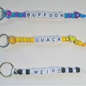 May include: Three keychains with different words spelled out in white letter beads. The first keychain has the word "BUFFOON" spelled out in white beads with blue and purple beads. The second keychain has the word "QUACK" spelled out in white beads with yellow beads. The third keychain has the word "WEIRD" spelled out in white beads with black and white beads.