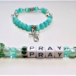 May include: A teal blue beaded bracelet with a silver cross charm and a teal blue beaded keychain with a silver charm and white letter beads spelling "PRAY PRAY".