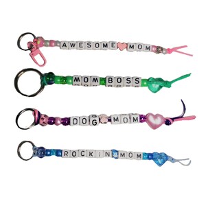 May include: Four keychains with colorful beads and letter tiles spelling out "Awesome Mom", "Mom Boss", "Dog Mom", and "Rockin Mom". Each keychain has a heart charm and a loop for attaching keys.