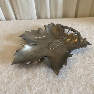 May include: A pewter-toned decorative leaf-shaped dish with a cluster of grapes and a vine detail. The dish has a detailed, textured surface and is likely made of metal. The design suggests a wine or vineyard theme.