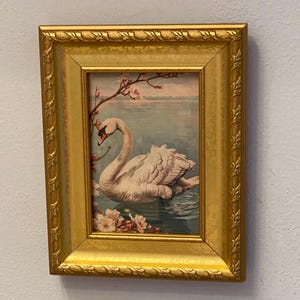 May include: A framed print featuring a swan on water, with pink flowers and branches. The artwork is set in a gold-colored frame with ornate detailing. The swan has a white body and a curved neck.