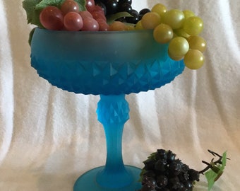 May include: A blue glass pedestal bowl with a diamond pattern, filled with green, red, and black grapes.