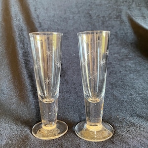 May include: Two clear glass shot glasses with a starburst design etched into the glass. The glasses have a flared top, a narrow stem, and a round base. The shot glasses are displayed on a dark blue surface.