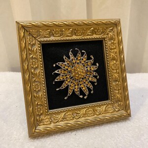 May include: A gold-framed display featuring a sunflower brooch. The brooch is silver-toned with a central cluster of yellow gemstones and radiating petals, also adorned with yellow gemstones. The frame has ornate floral detailing and a black velvet backing.