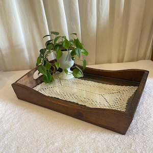 May include: A rectangular dark brown wooden serving tray with cutout handles on the sides. The base of the tray features a cream-colored rectangular crochet doily under a glass surface. A small green plant sits in a white fluted vase on the tray.