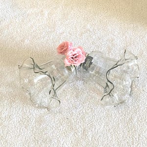 May include: Two clear, decorative glass vases with ruffled edges are connected, holding several pink roses. The vases are on a white, textured surface, creating a delicate and elegant floral arrangement.