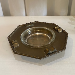 May include: A vintage brass ashtray with a clear glass insert. The ashtray has a hexagonal shape with decorative edges and three built-in cigarette rests. The brass has a warm, aged patina.