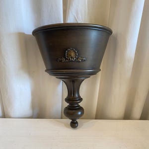 May include: A dark brown wall-mounted planter with a semi-circular basin and a tapered, pedestal-style base. The front of the basin features a centered, ornate gold-toned metal embellishment shaped like a shell with decorative scrollwork.