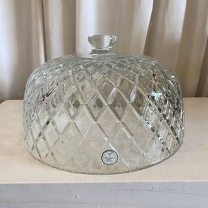 May include: A clear glass dome food cover featuring a raised diamond pattern across its surface and a small circular knob handle at the top. A small round sticker with the text Crystal Clear is visible on the lower front of the glass dome.