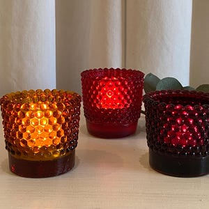 May include: Three glass votive candle holders in amber, red, and burgundy. Each holder has a textured, bubble-like surface. The amber votive is lit with a warm glow, while the red and burgundy ones emit a red light.