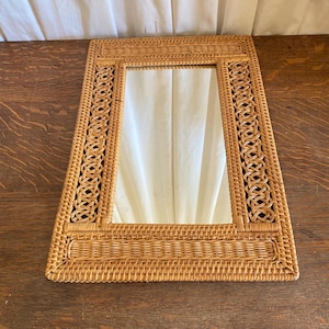 May include: A rectangular wall mirror featuring a light brown woven rattan frame. The frame displays a combination of intricate circular lattice patterns along the sides and a tight, horizontal weave along the top and bottom edges.