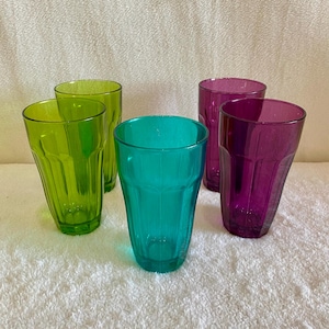 May include: Five colorful drinking glasses in shades of green, teal, and purple. The glasses have a faceted design and are made of transparent material. The glasses are arranged on a white surface.