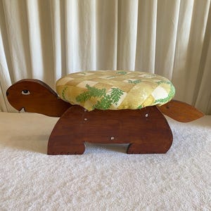 May include: A wooden turtle-shaped footstool with a padded seat. The seat has a yellow and green patterned fabric. The turtle's body is made of dark brown wood, with the head, legs, and tail extending from the body.