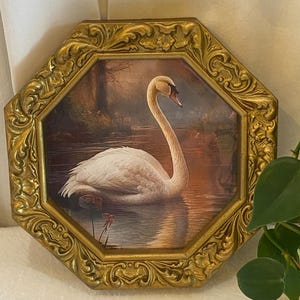 May include: An octagonal framed painting featuring a white swan gracefully gliding on water. The artwork is set within an ornate gold-colored frame with detailed scrollwork. The swan has a long, curved neck and a red beak. The background shows a serene landscape.